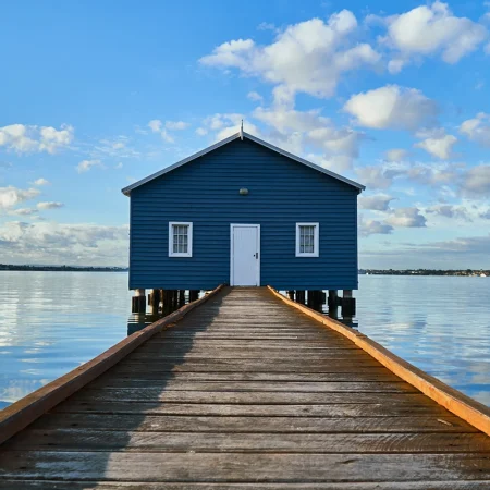 Blue boathouse at the end of a pier over a lake with a blue cloudy sky.