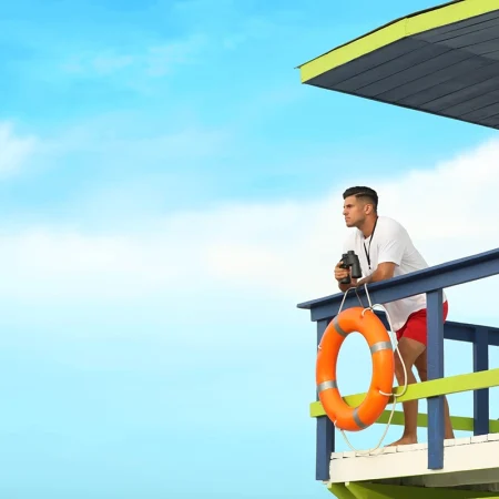 Male lifeguard on a tower looking out and holding binoculars.